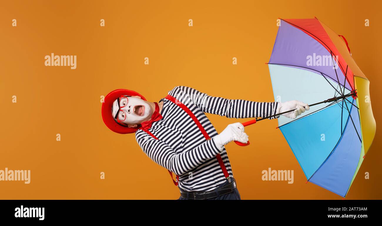 Mime man with multi-colored umbrella during wind isolated on empty orange background in studio. Stock Photo