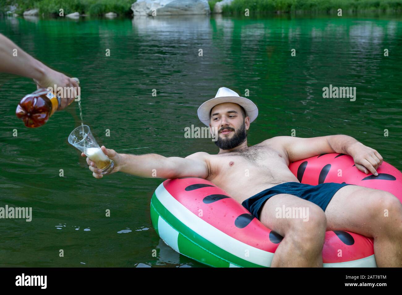 young hipster bearded guy, man in hat lying or relaxing on inflatable circle, beer is poured into his outstretched hand with glass Stock Photo