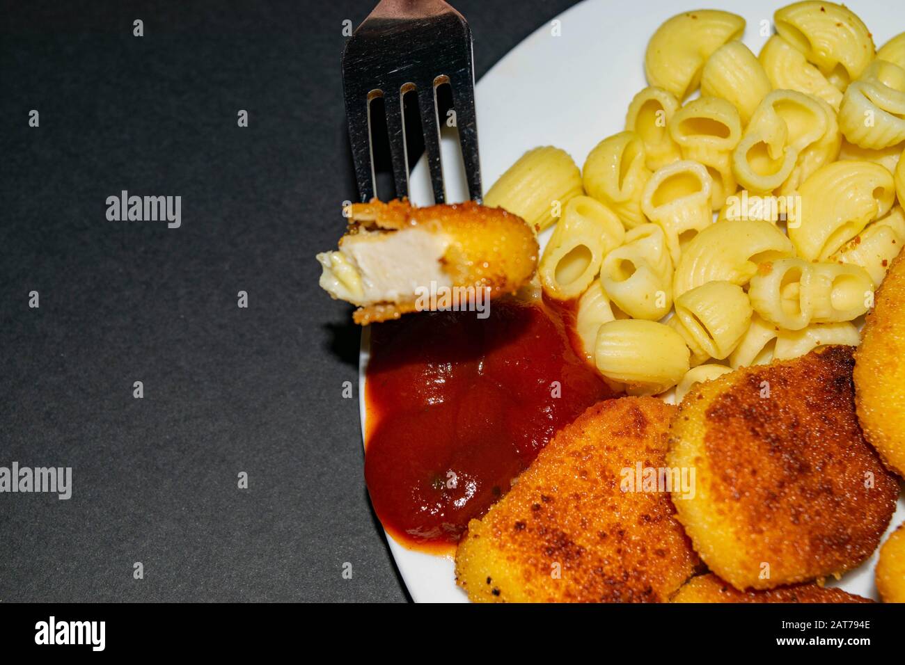 A piece of nuggets on a fork and a white plate with fried chicken nuggets, pasta and ketchup on a dark background. Close up Stock Photo