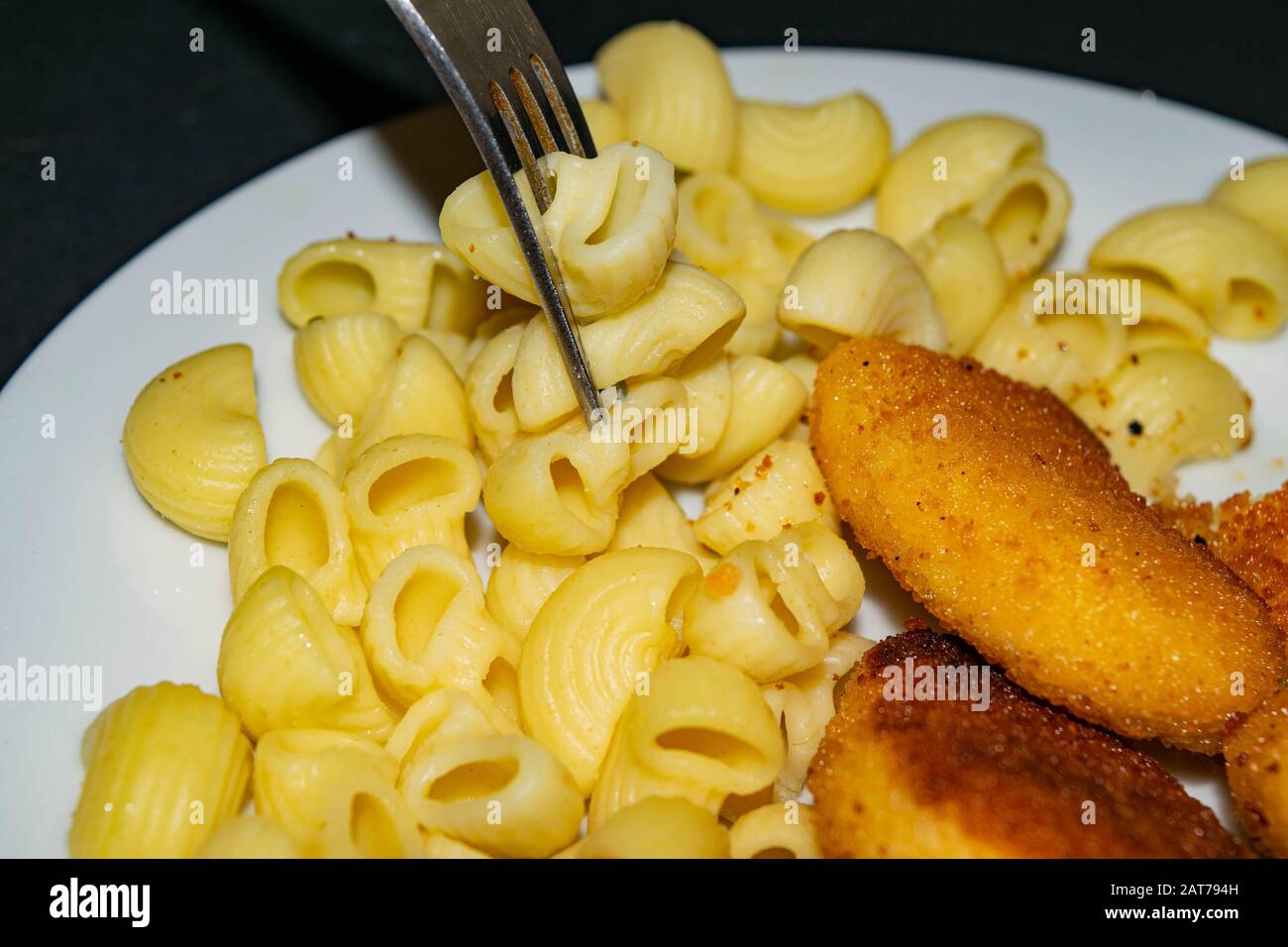 A few boiled macaroni on a fork and a white plate with fried chicken nuggets, macaroni and ketchup on a dark background. Close up Stock Photo