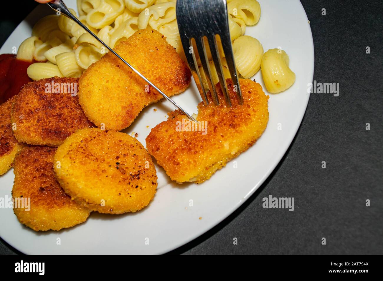 Fried chicken nuggets and boiled pasta with ketchup on a white plate on a dark background. Fork and knife cutting up the nuggets. Close up Stock Photo