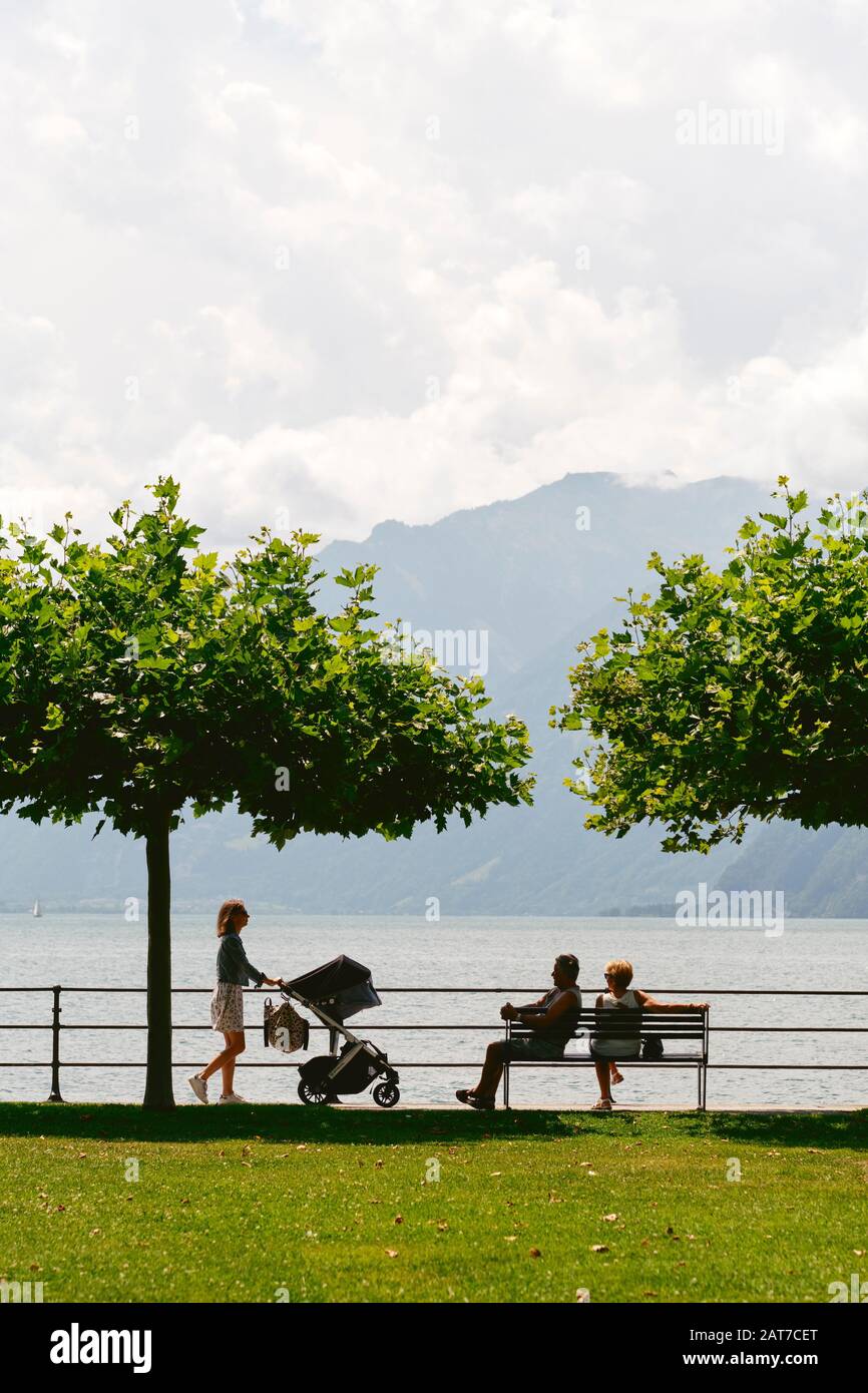 A young woman with a pram and a retired couple sitting on a bench overlooking Lake Lucerne in Brunnen, Schwyz, Switzerland EU Stock Photo