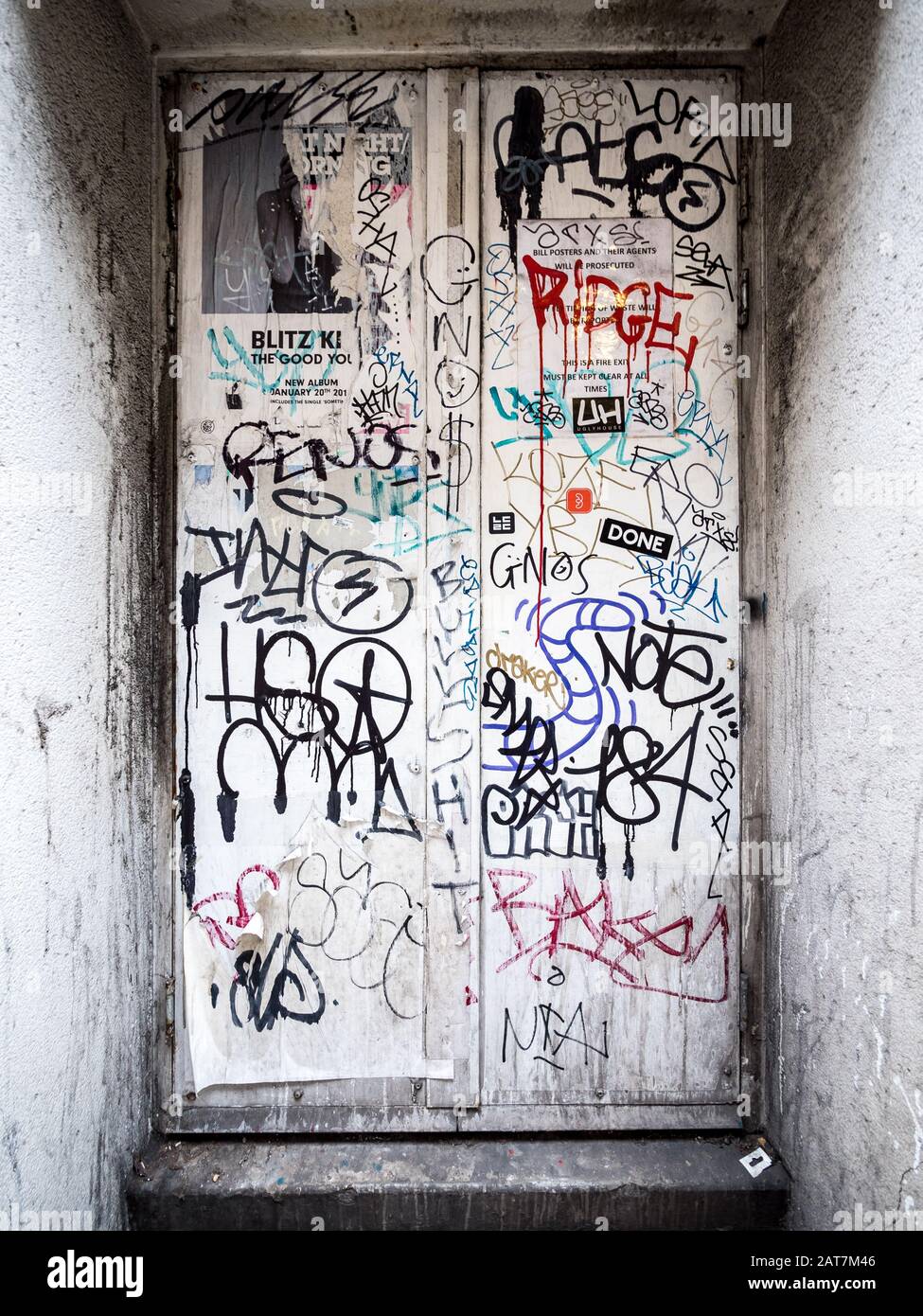Urban Anarchy. Detail of a recessed white door covered in graffiti tags defying a defaced warning sign. Stock Photo
