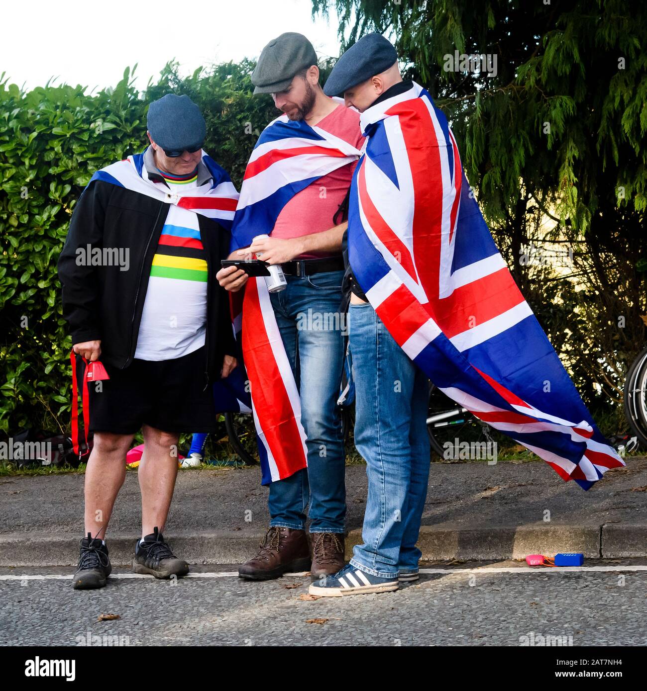3 men looking at phone, standing at side of road, wearing flat caps & Union Jack flags, spectators at sports event - Harrogate, Yorkshire, England UK Stock Photo
