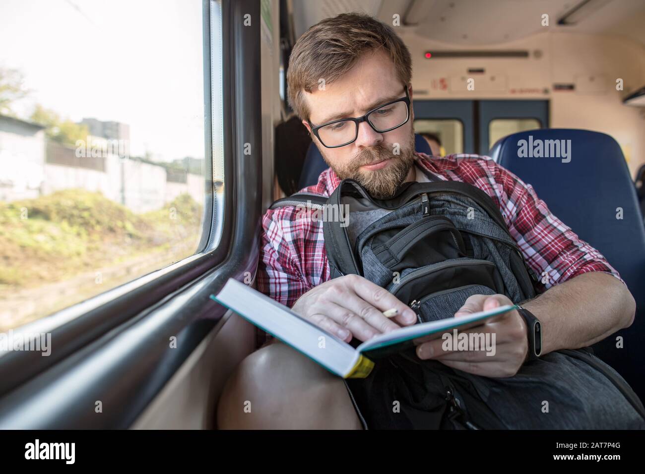 Focused bearded man riding in a train, reading a book and making notes with a pencil. Stock Photo