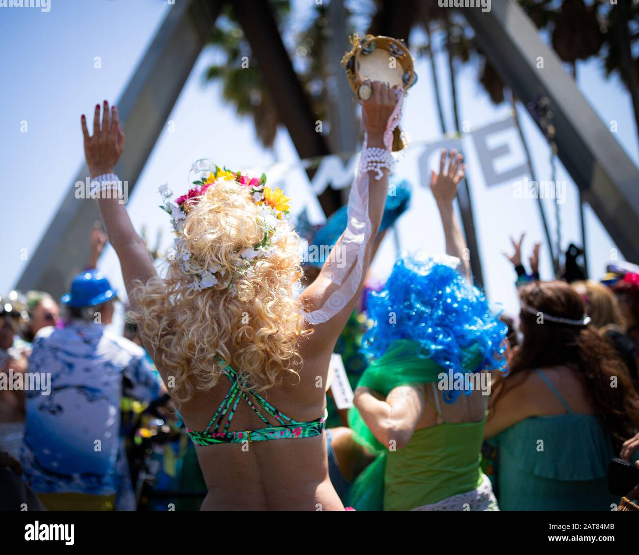 Festive Parade in Venice Beach, California with Blonde Female in Bikini Top Holding Up Arms and is holding a tambourine in one hand. Stock Photo