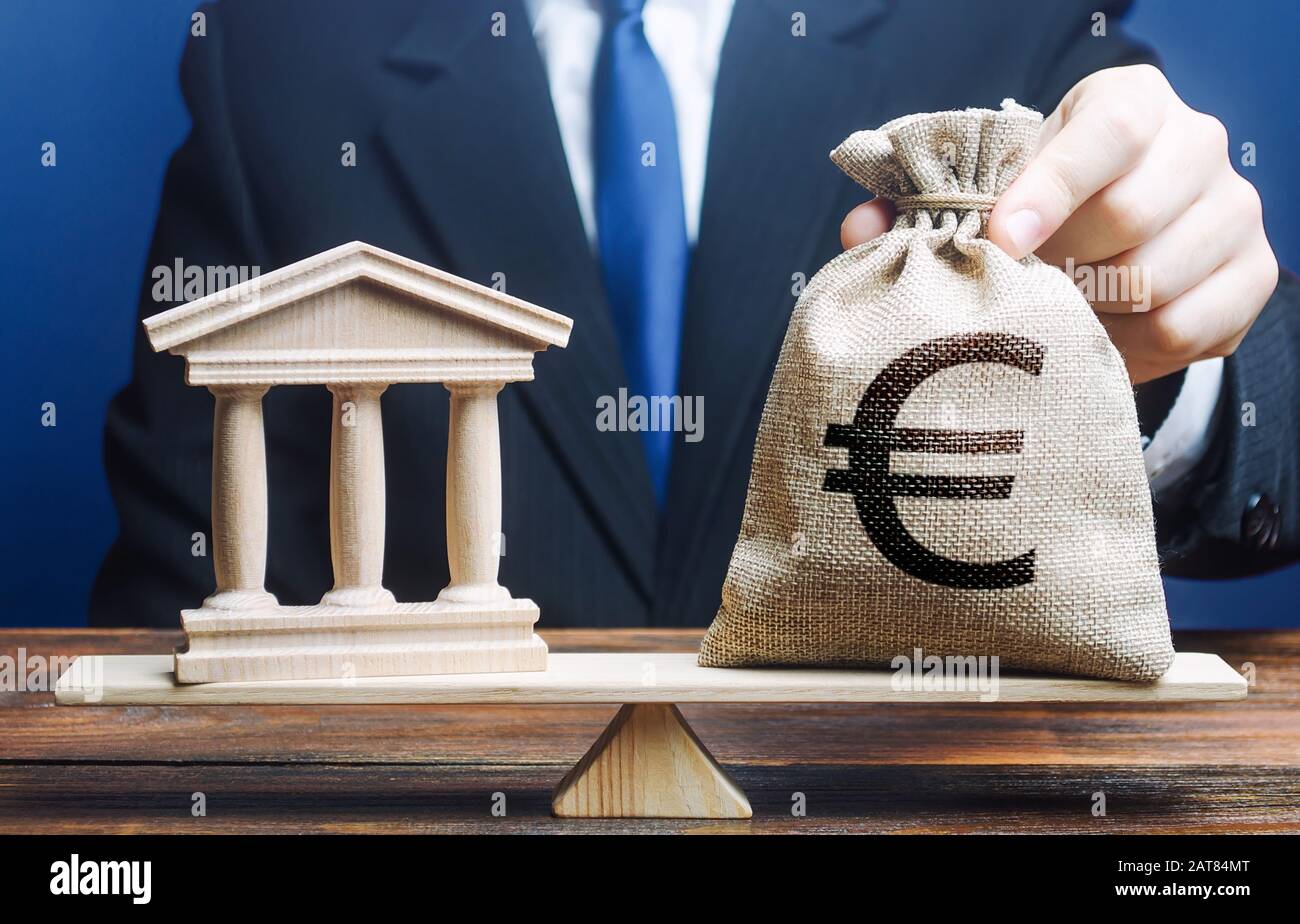 A man puts a euro money bag on scales opposite to government building, bank, university. Taxes payment. Lobbying interests. Deposits, loans. Financial Stock Photo