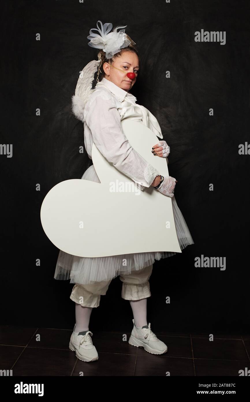 Serious woman in clown costume holding white empty heart banner on black background Stock Photo