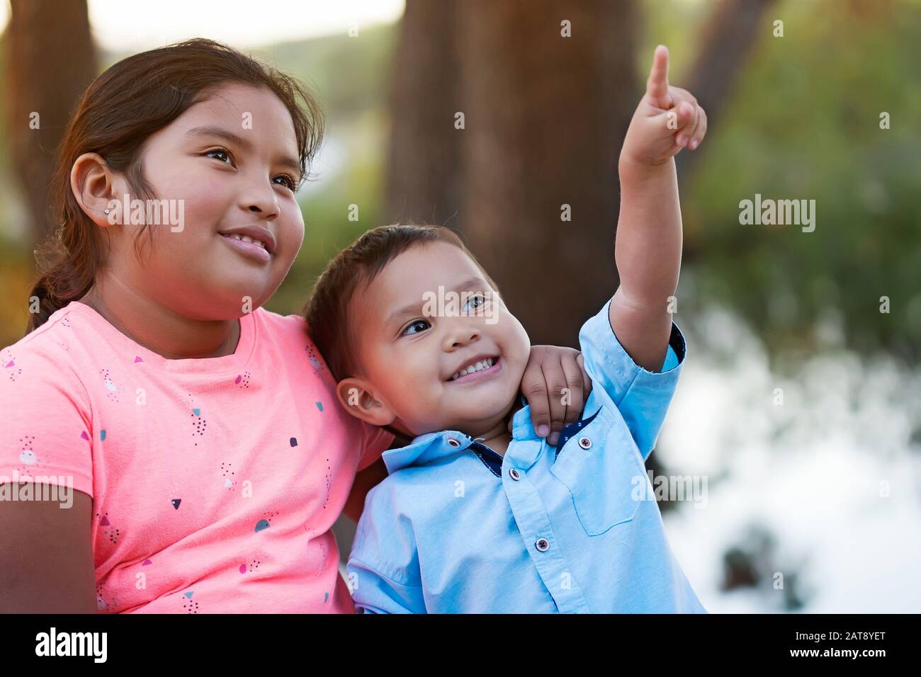 A happy younger brother looks and points to the sky while his older sister puts her arm around his neck and smiles. Stock Photo