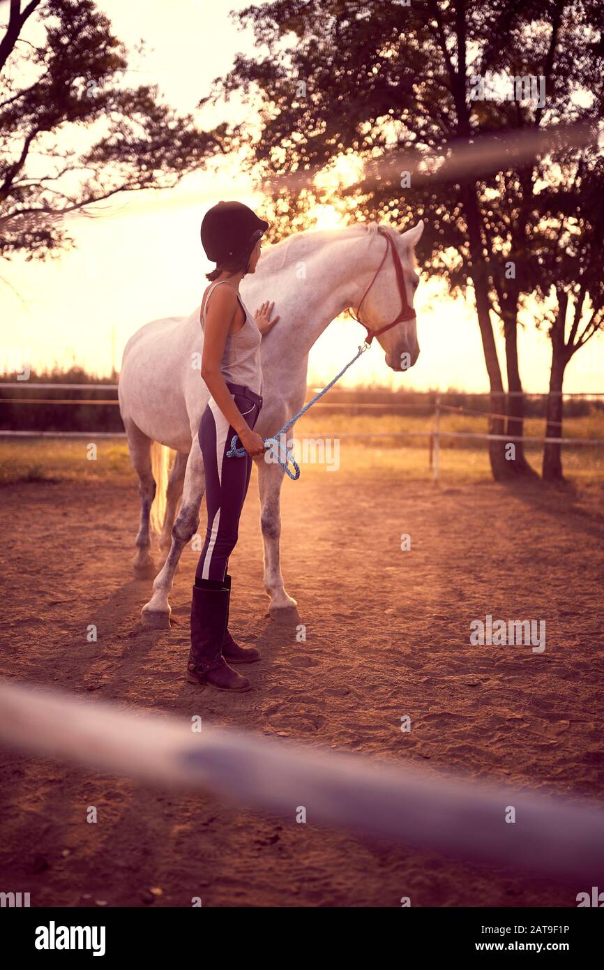 Woman in equestrian gear petting a horse while looking away at the sunset. fun on countryside,  golden hour. Freedom nature concept. Stock Photo