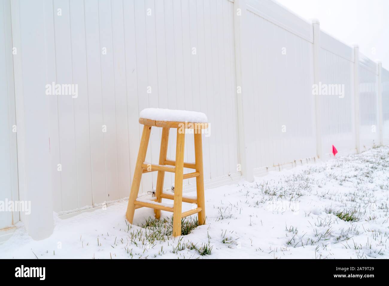 Wooden stool on snow covered ground in winter against white wood fence Stock Photo
