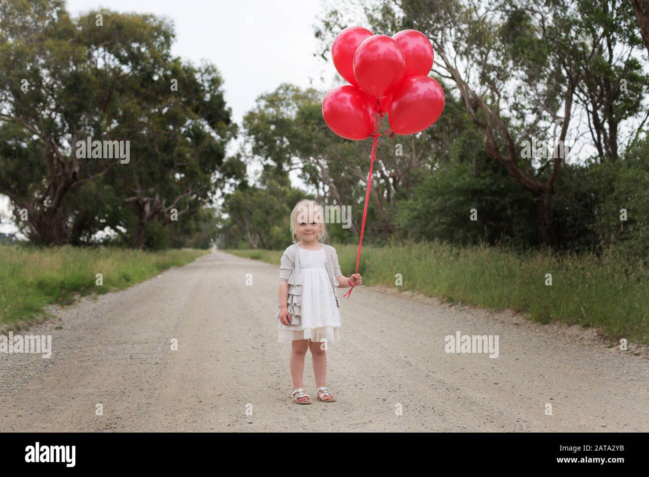 Australian Caucasian girl standing on a dirt road in the country holding red balloons Stock Photo