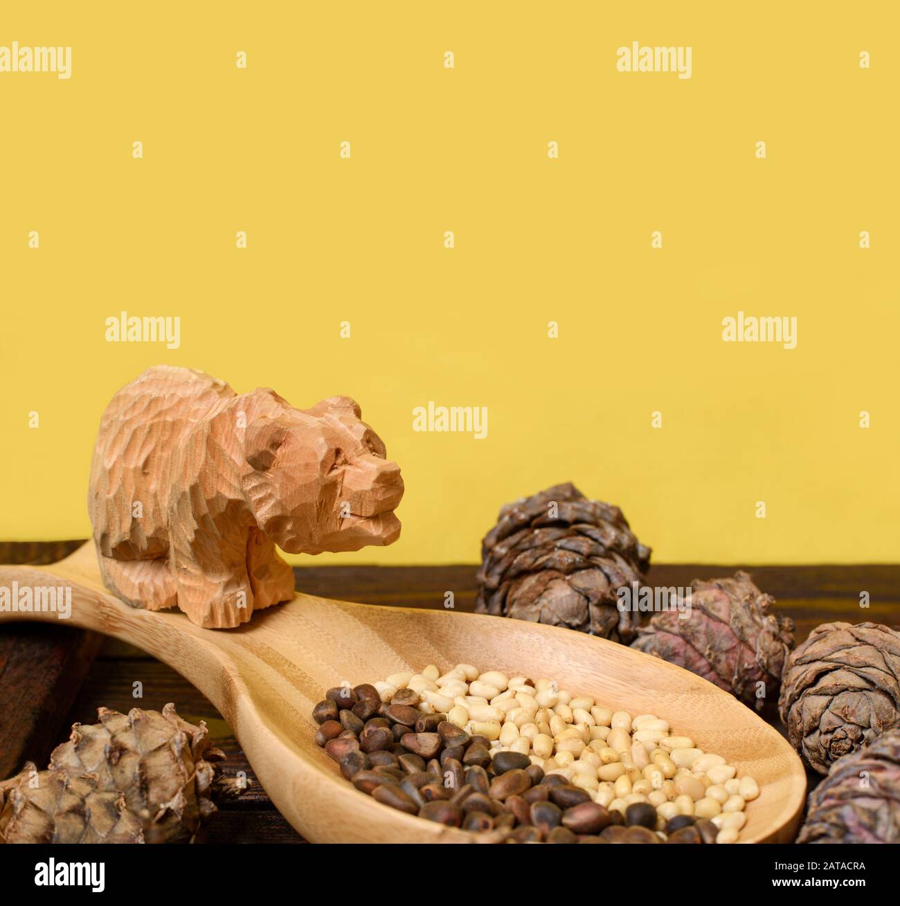 Pine nuts. Cedar nuts and cones on wooden table. Wooden bear made of cedar on yellow background. Кедровые орешки. Кедровые шишки. Деревянный медведь. Stock Photo