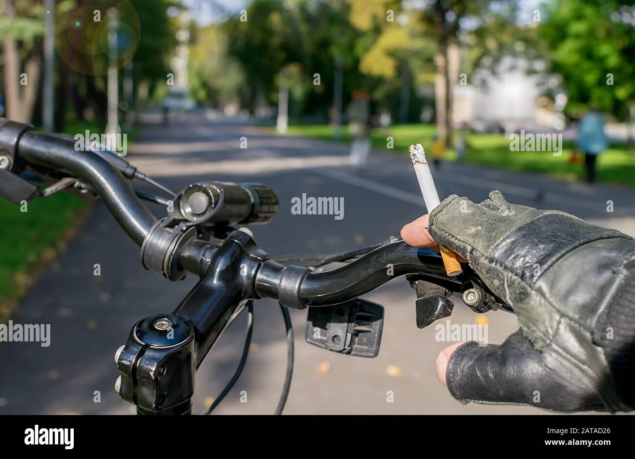 cigarette in the hand of a smoking cyclist who rides a bike on the  background of the asphalted path of the city park Stock Photo