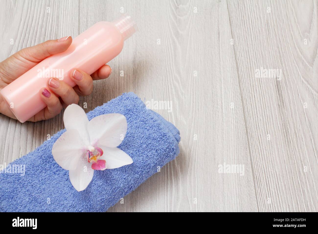 Bottles of shampoo in woman's hand, towel with orchid on a gray wooden background with copy space. Women cosmetics and accessories. Top view. Stock Photo