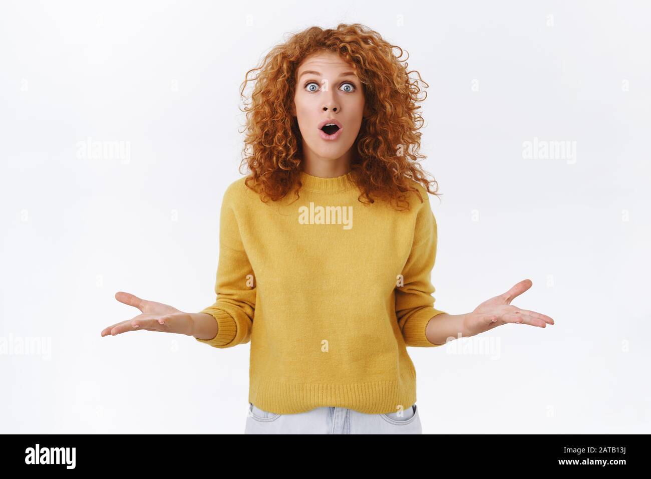 Shocked, speechless and amazed redhead curly woman in yellow sweater losing speech from astonishment and shock, raise arms sideways, gasping and open Stock Photo