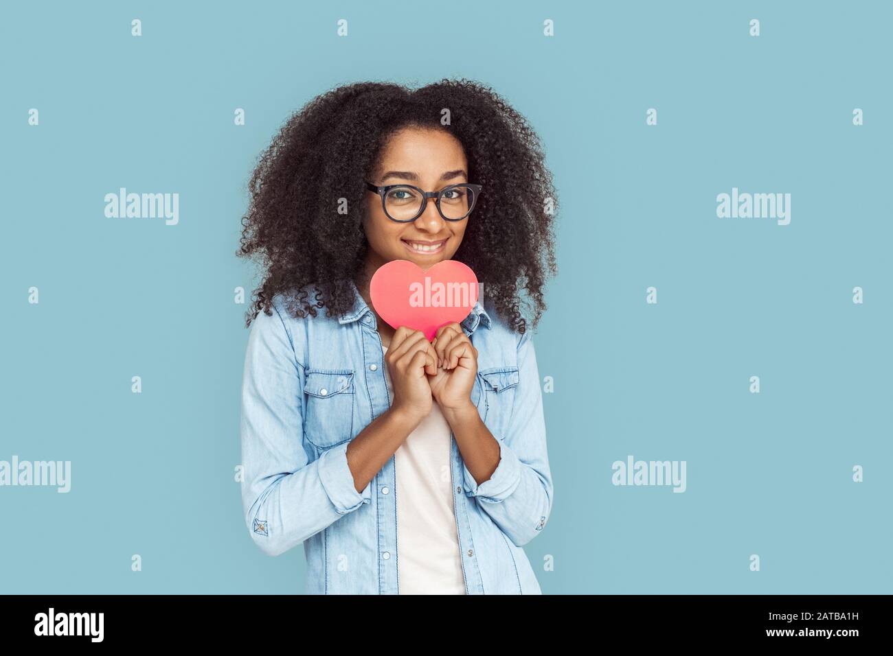 Freestyle. African girl in glasses standing isolated on gray with heart card biting lip playful Stock Photo