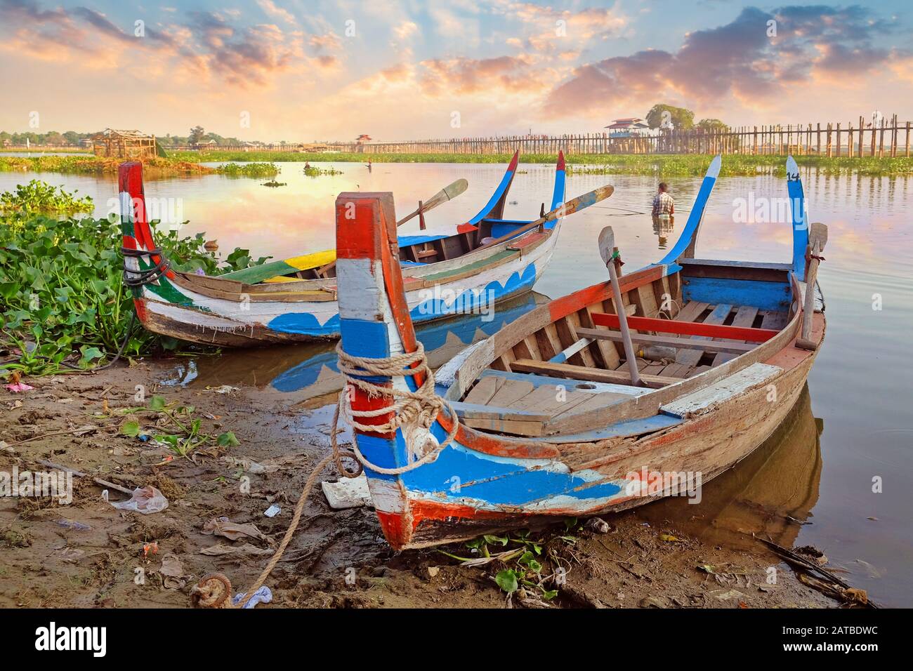 Colorful traditional boats on the side of the Taung Tha Man Lake, near Mandalay, Myanmar, surrounded by green vegetation, against a beautiful colorful Stock Photo