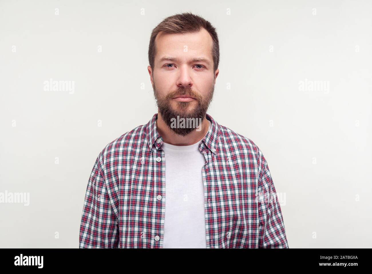 Portrait of serious unhappy despondent young man with beard in plaid shirt looking at camera with no enthusiasm, calm gloomy expression, bad mood. ind Stock Photo