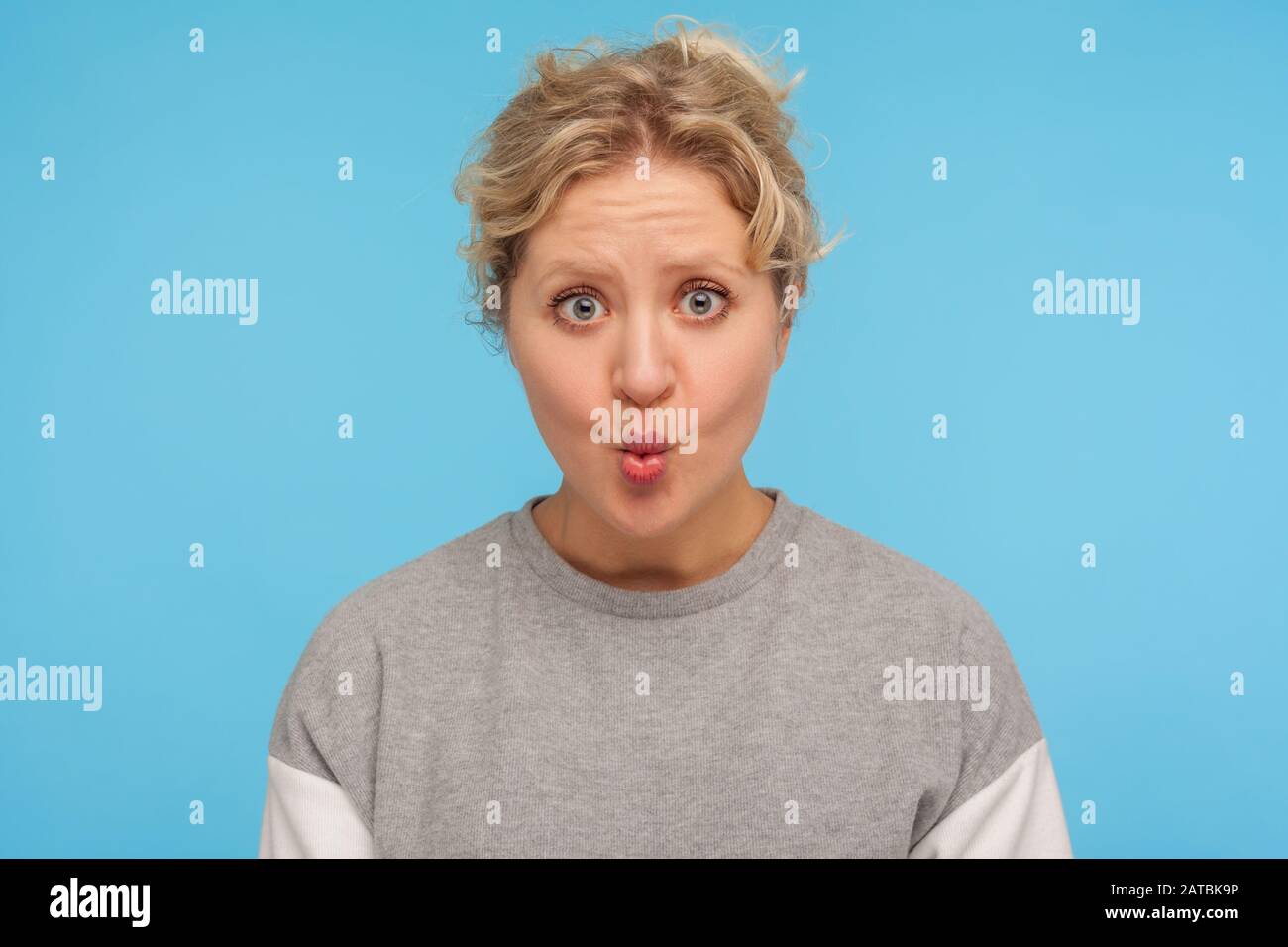 Funny woman with short hair in casual grey sweatshirt showing fish face with pout lips and big eyes, astonished surprised expression, making faces. in Stock Photo