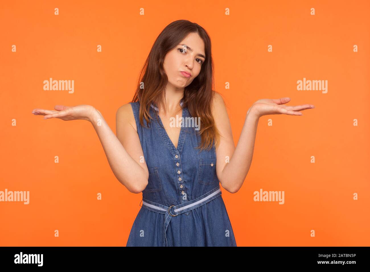 I don't know! Confused brunette woman in denim dress showing indifference and embarrassment with raised hands and clueless doubtful face expression, h Stock Photo