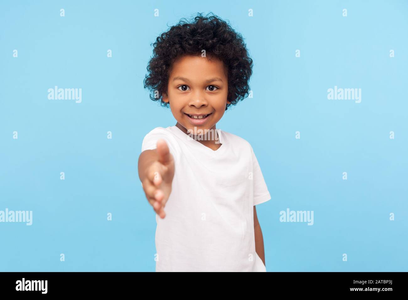 Portrait of friendly cheerful little boy with curly hair in white T-shirt giving hand to handshake and smiling at camera, looking gullible trustful. i Stock Photo