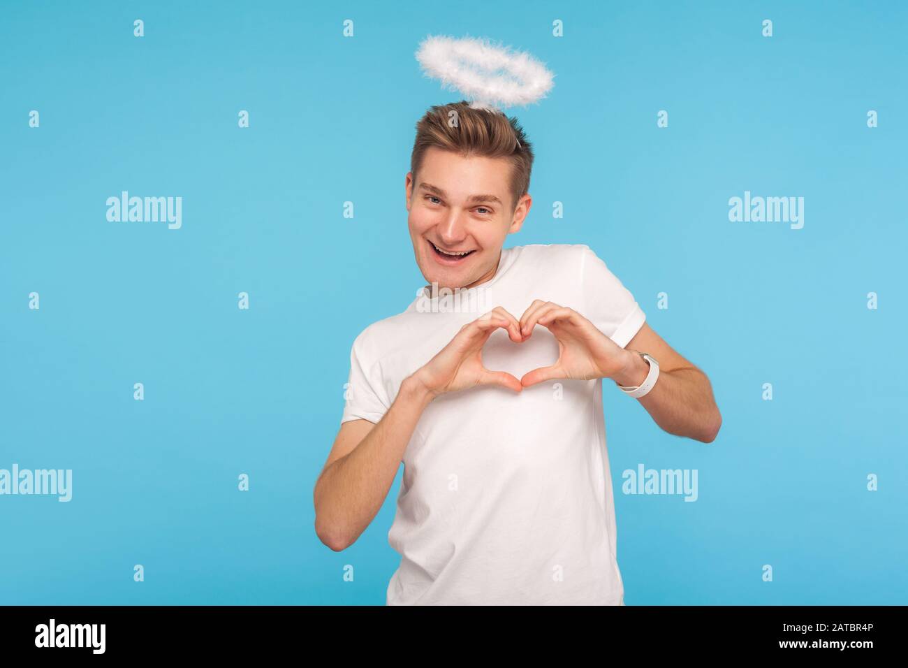 Portrait of happy angelic man with halo above head making heart shape with hands and smiling to camera, sharing romantic amorous feelings, declaration Stock Photo