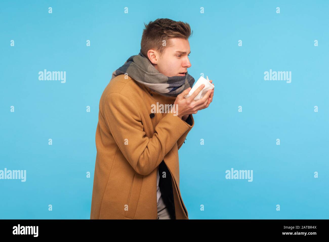 Influenza season. Portrait of flu-sick man in autumn coat and scarf sneezing in napkin, feeling unwell with runny nose, caught cold or allergy symptom Stock Photo
