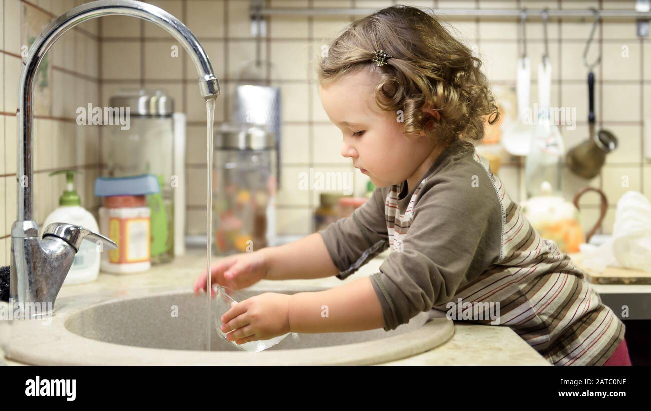 Little child is washing dishes in kitchen. Nice toddler learns the