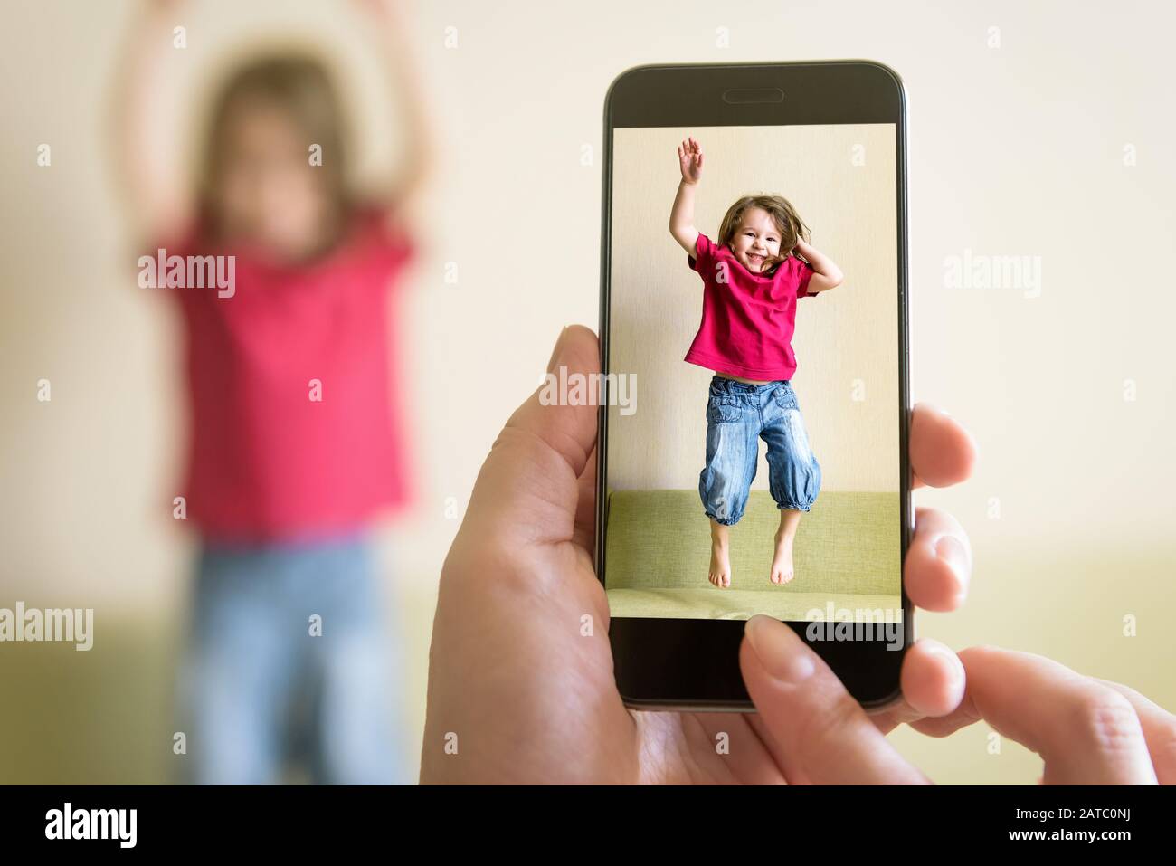 Funny three-year-old child jump on couch. Mother taking photo of baby girl with her mobile phone. Photography of happy kid playing and jumping at home Stock Photo