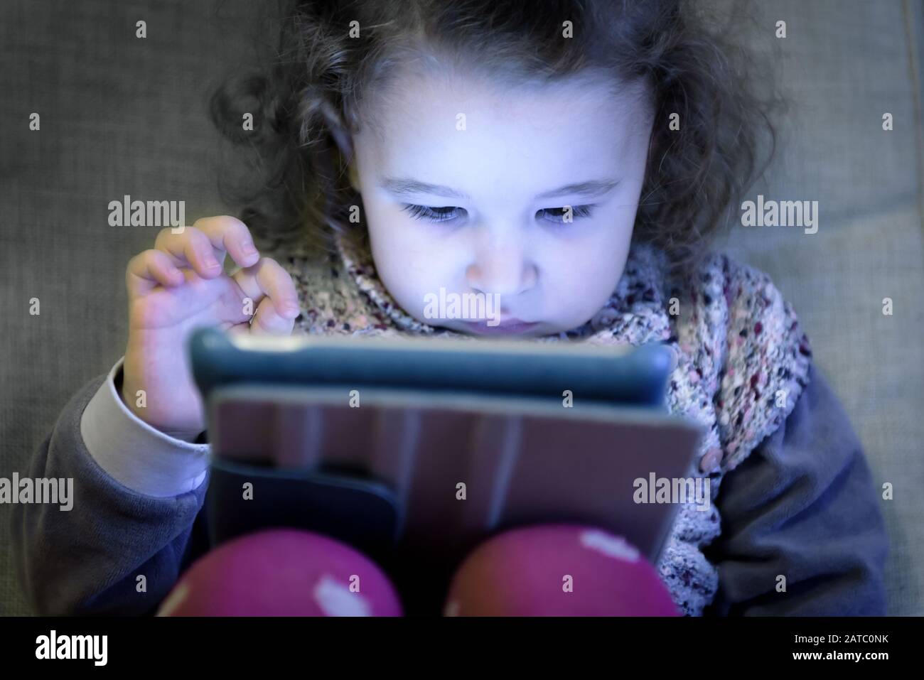 Little girl uses a digital tablet. Two-year-old child looks electronic device at home. Cute toddler sits on a couch and plays computer games. Face of Stock Photo