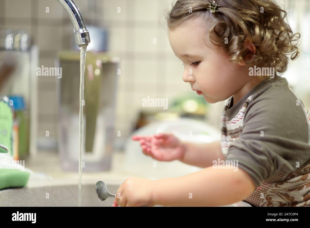 Pretty kid is washing dishes in kitchen. Nice toddler learns the household. Beautiful two-year girl washes spoon in sink. Adorable child does house cl Stock Photo