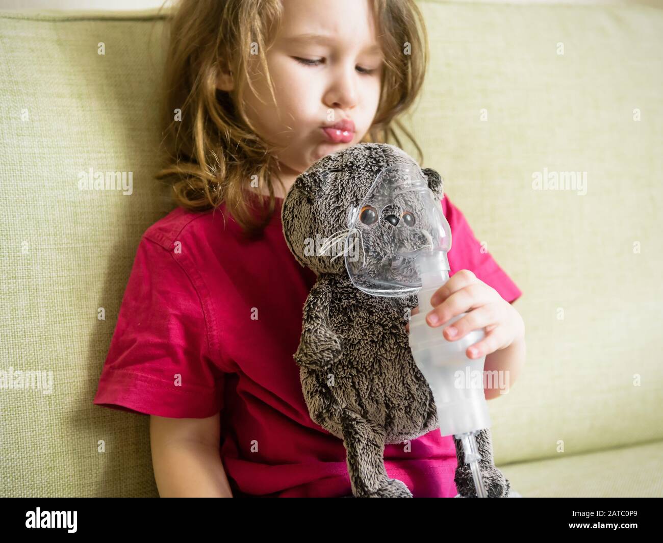 Little girl holds inhaler mask at home. Funny kid plays with toy and nebulizer. Baby using equipment to treat asthma or bronchitis. Concept of treatme Stock Photo