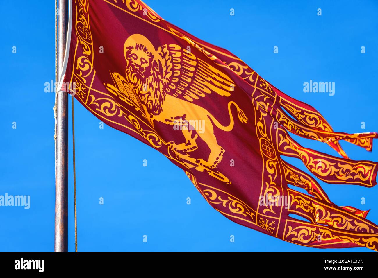 Flag of Venice waving on street in Venice, Italy. Golden winged lion on red flag, closeup view. Old Venetian flag is on background of blue sky in summ Stock Photo