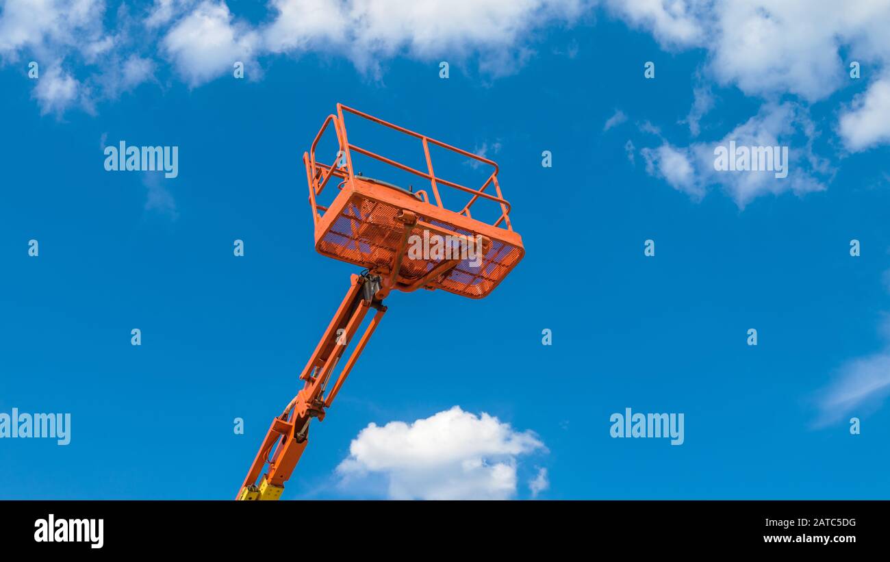 Cherry picker on blue sky background. Boom with lift bucket of heavy machinery. Orange platform of the telescopic construction lift in summer. Stock Photo