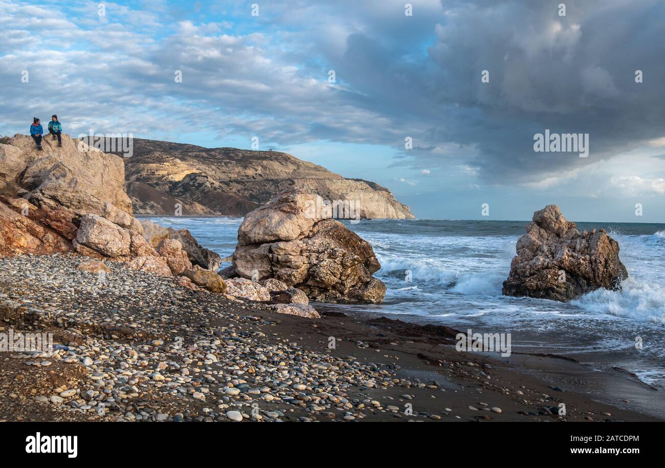 Two tourist women dressed in winter clothing sitting on a rock enjoying the  stormy seascape. Rock of Aphrodite in Cyprus Stock Photo