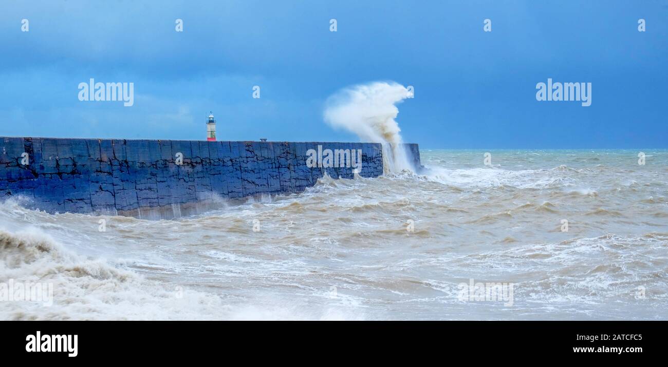A harbor wall with a rough stormy sea crashing against the wall causing the sea to be blurred and in motion, behind is a red and white lighthouse, wav Stock Photo