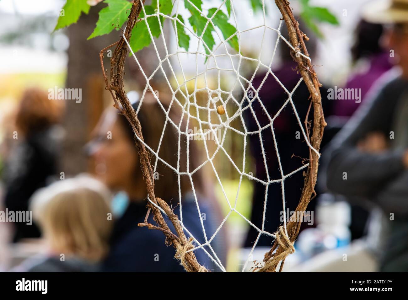 Closeup of handmade dreamcatcher hanging on tree branch with defocus group of people sitting and looking at something at world and spoken word event Stock Photo