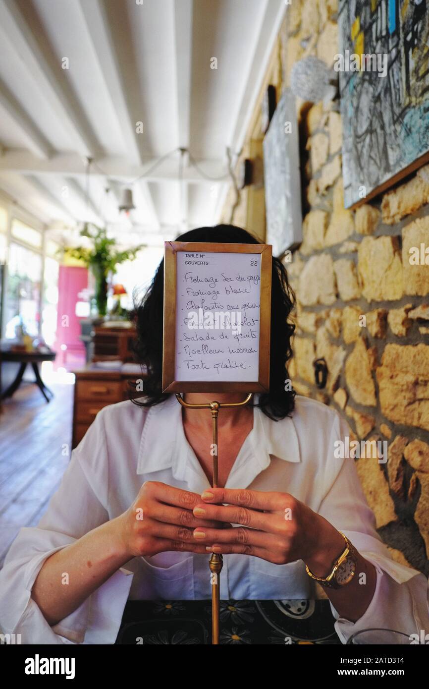 A woman sits at table in a French bistro, village of Oingt; her hand hold the menu stand, the menu becomes trompe l'oeil in place of her face Stock Photo
