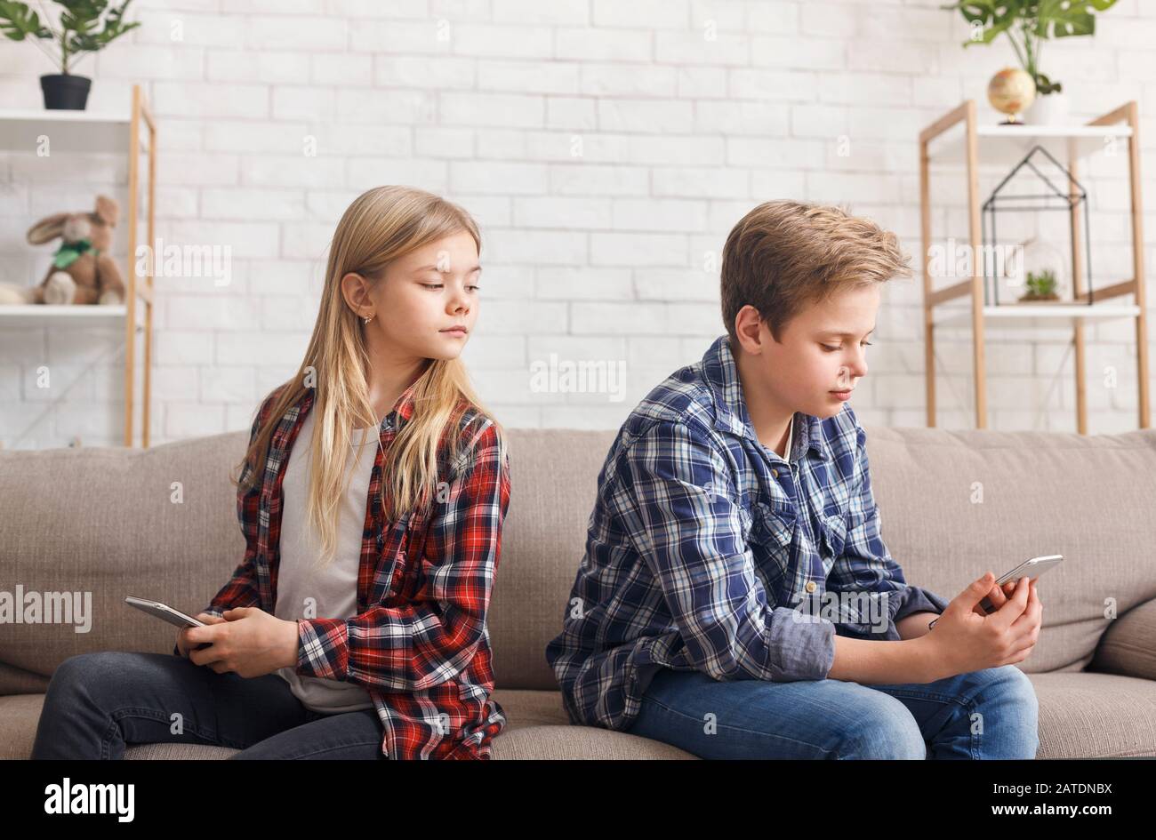 Girl Using Smartphone Peeking At Boy's Chat Sitting On Sofa Stock Photo