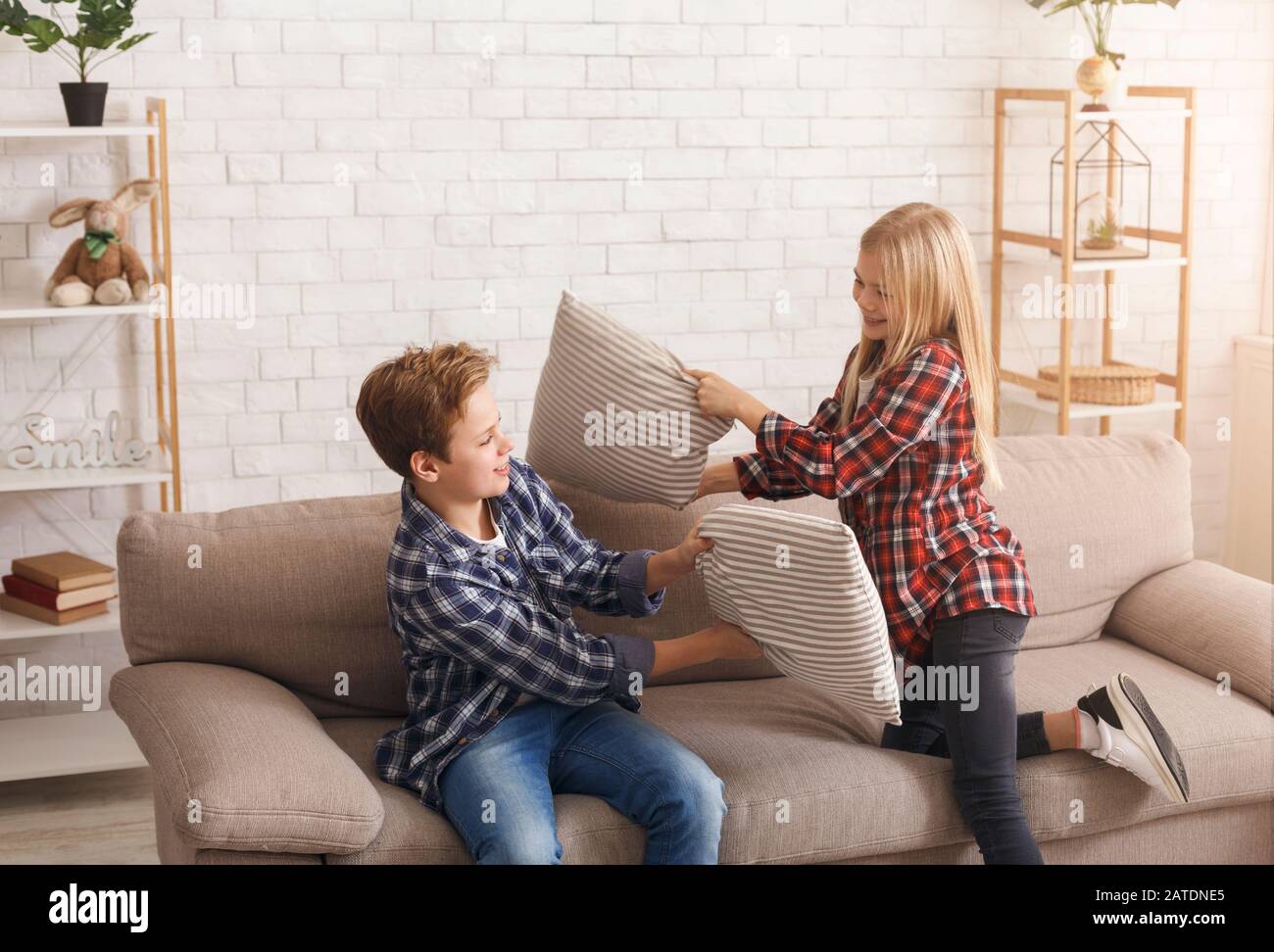 Happy Brother And Sister Having Pillow Fight On Couch Indoor Stock Photo