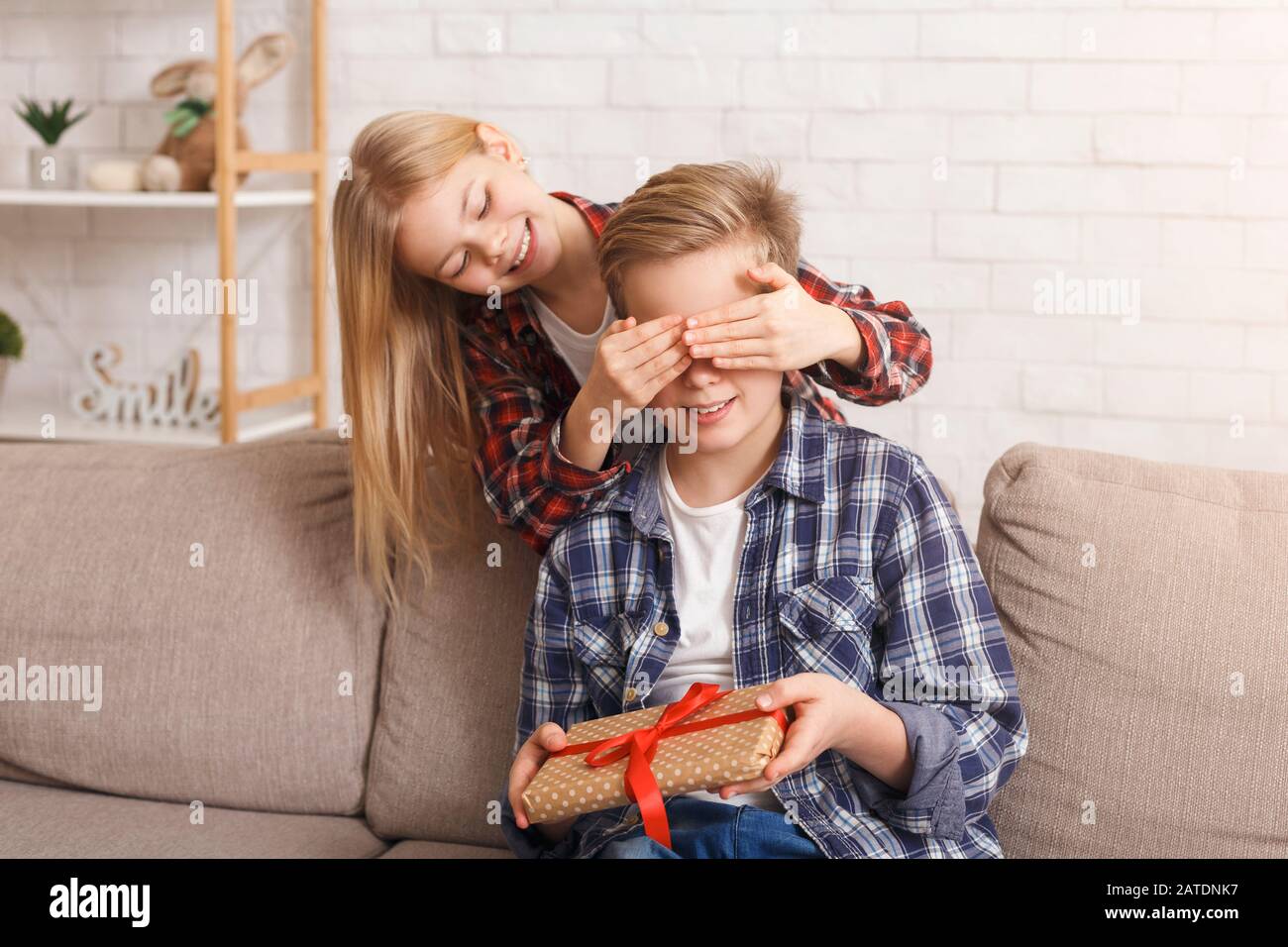 Sister Covering Brother's Eyes Giving Birthday Gift Sitting At Home Stock Photo