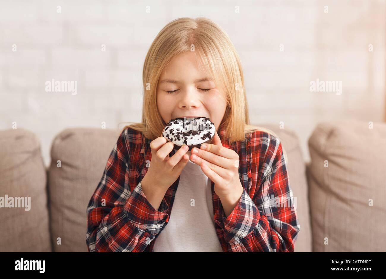 Little Girl Eating Donut Sitting On Couch At Home Stock Photo