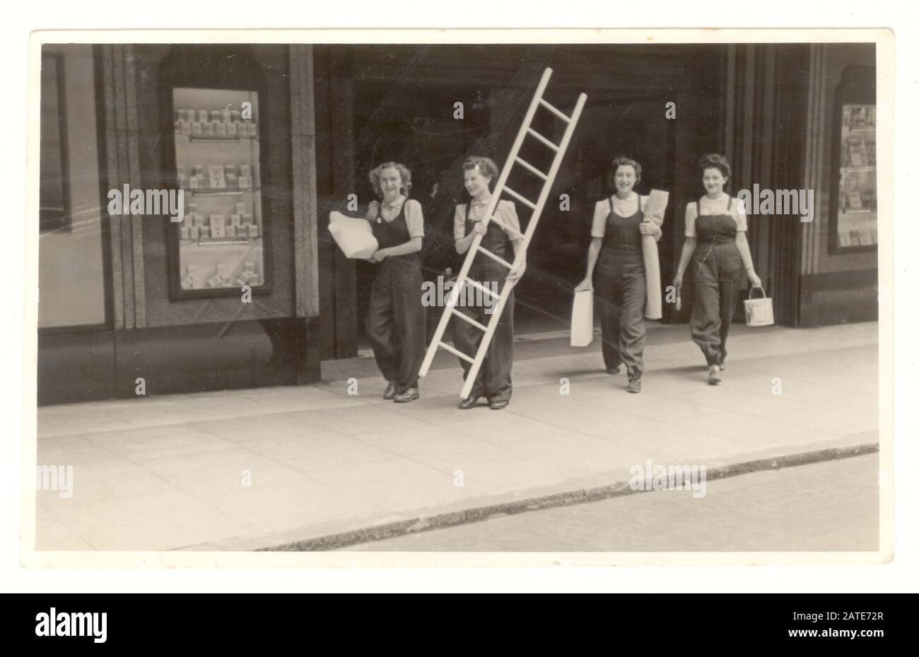 WW2 era photo of women employees, department store window dressers wearing dungarees carrying paint, fabric and a mannequin walking through the shop entrance, dated 1942, U.K. Stock Photo