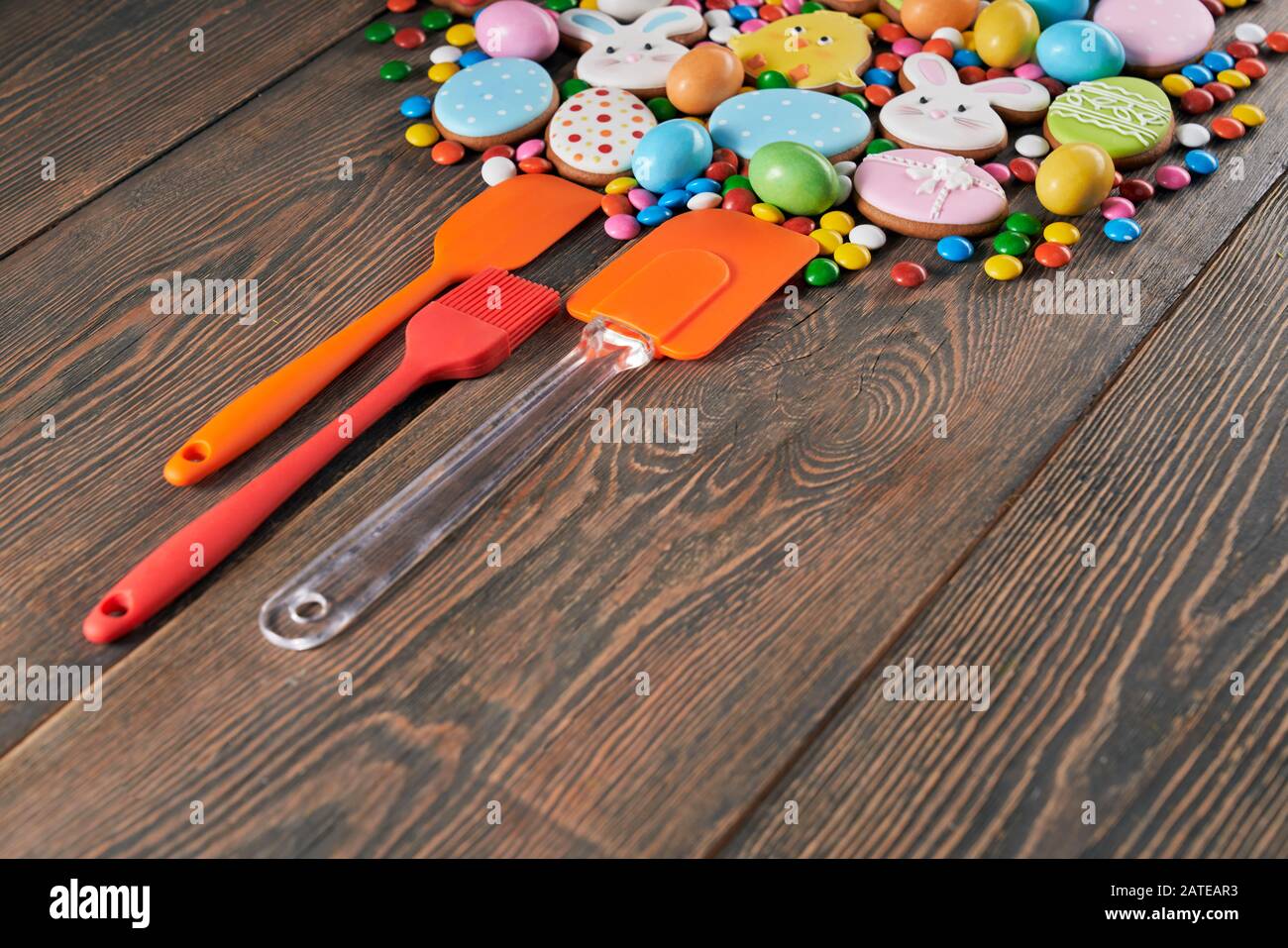 Crop of colorful ginger glazed cookies and chocolate balls with sugar shell isolated on wooden table. Homemade pastry in shape of easter animals, eggs and carrots, three silicone kitchen tools nearby. Stock Photo