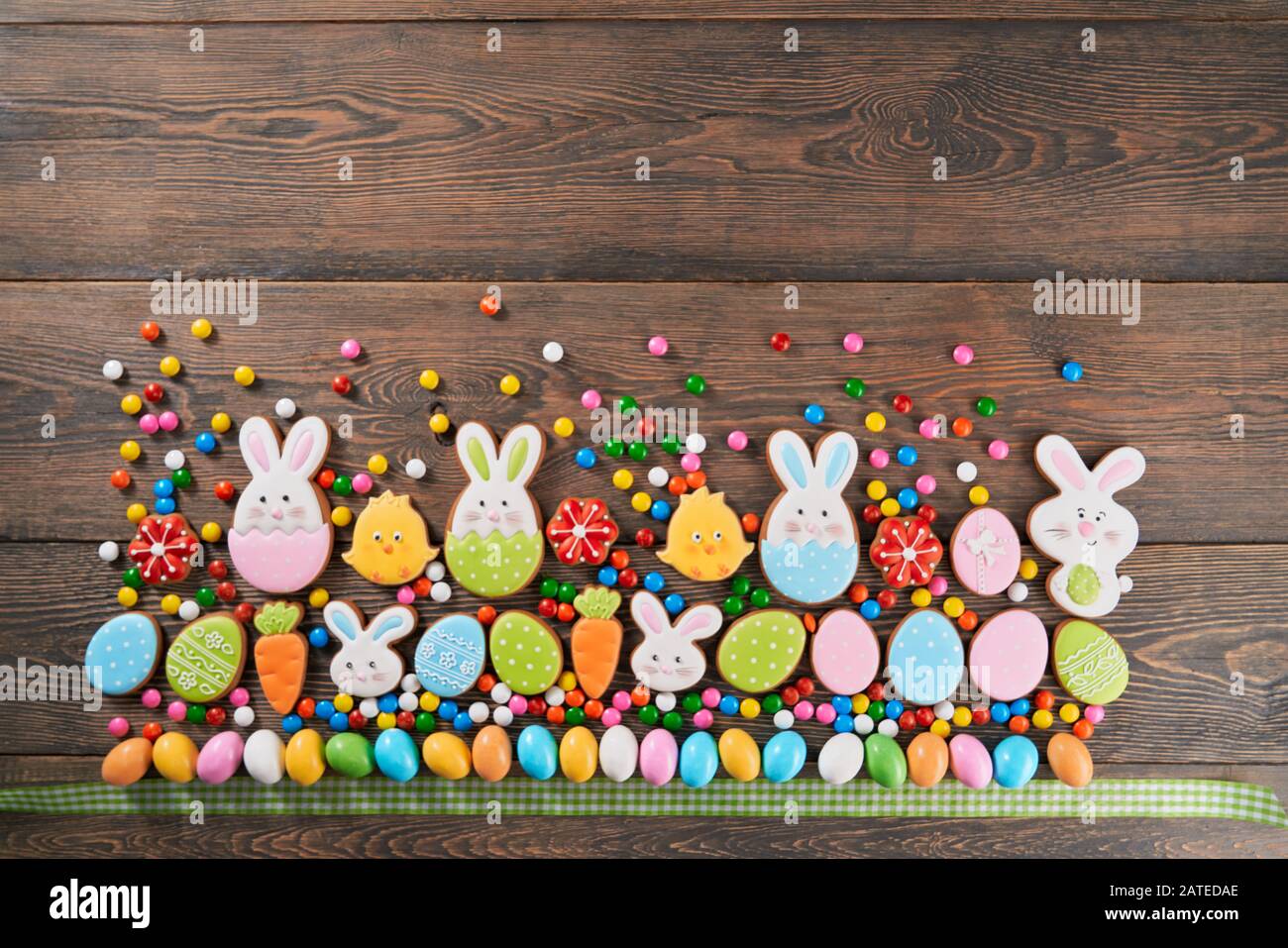 Big and small colorful peanut chocolate balls in crispy sugar shell and tartan ribbon isolated on wooden background. Easter ginger glazed cookies in shape of bunnies, flowers, carrots and eggs in row. Stock Photo