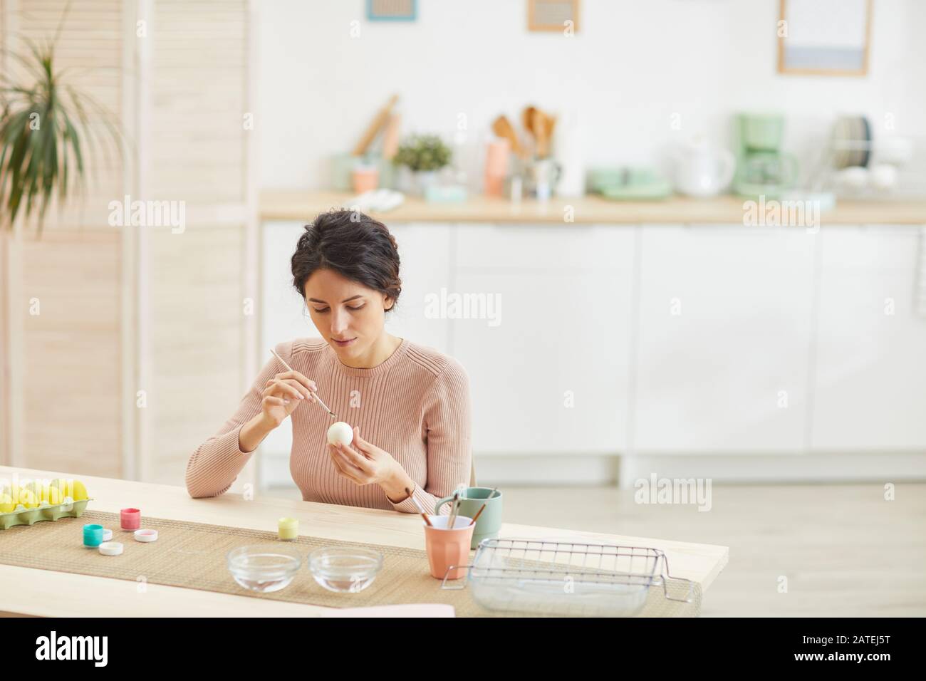 Wide angle portrait of adult woman painting Easter eggs in light kitchen interior, copy space Stock Photo