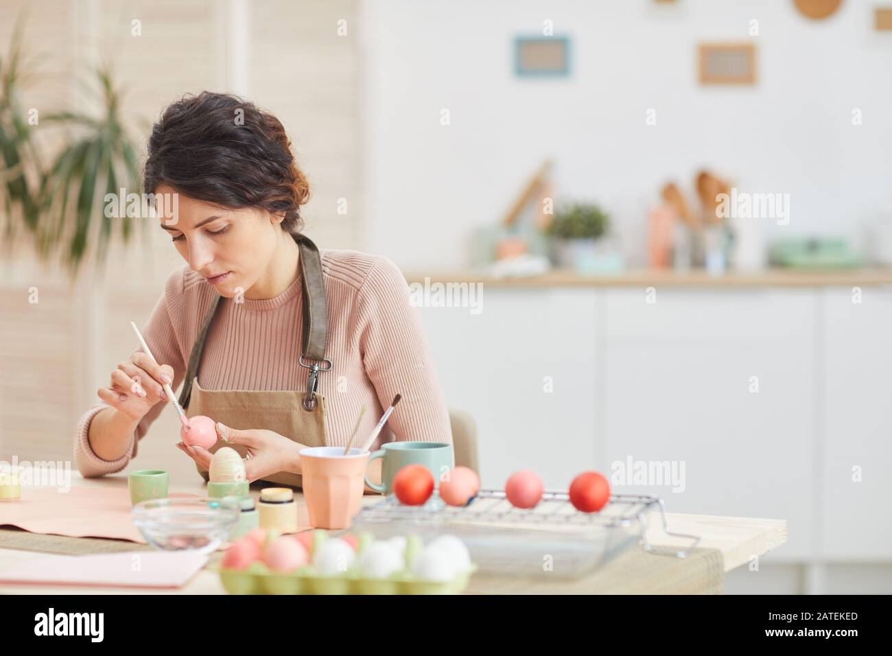 Portrait of adult woman painting eggs in pastel colors for Easter while sitting at table in cozy kitchen interior, copy space Stock Photo