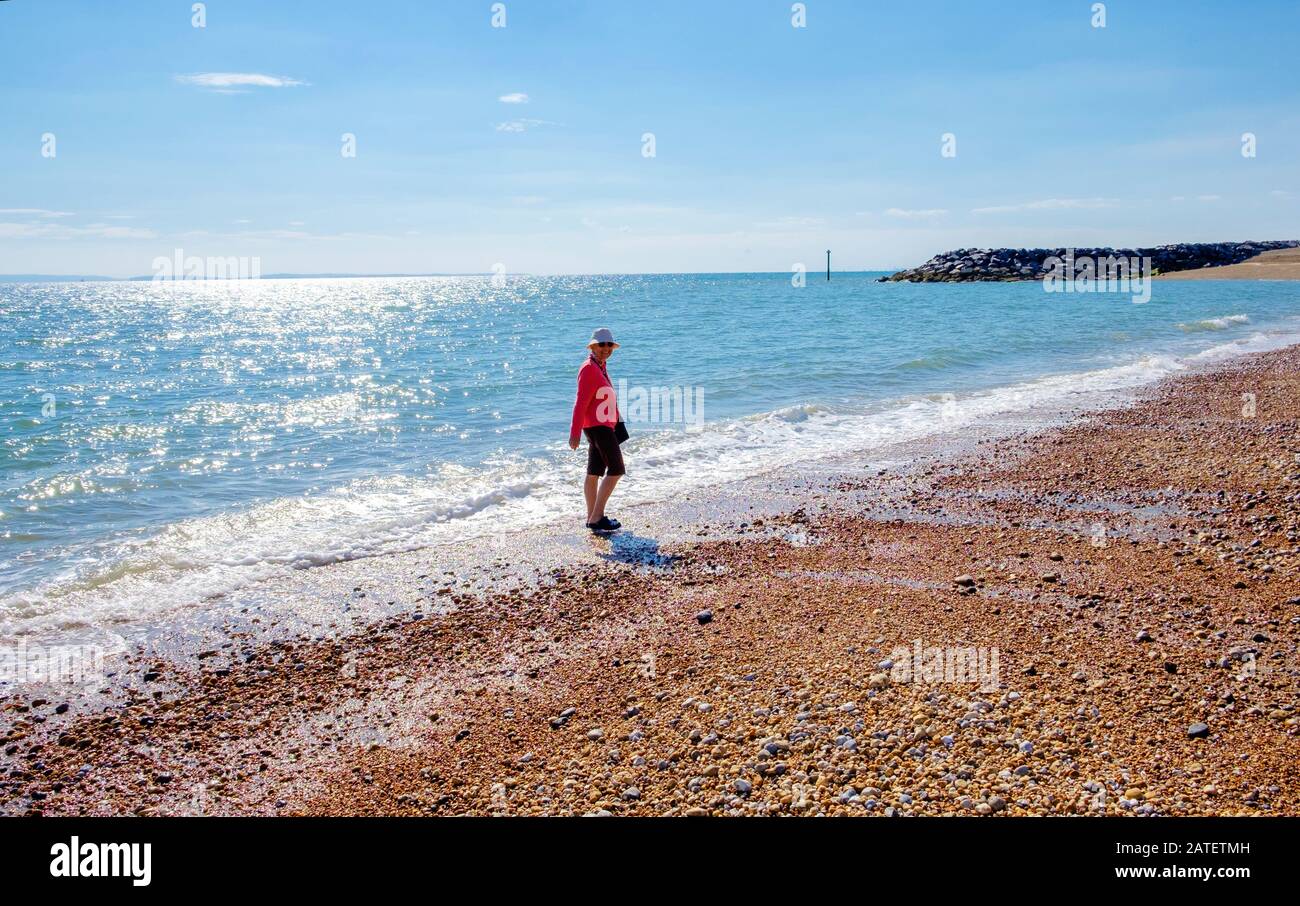 A lady smiles & walks along the water edge on Beautiful Beach, Selsey, West Sussex, England. Stone breakwater & Bracklesham Bay in background. Stock Photo