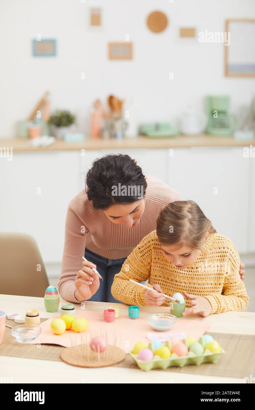 Vertical portrait of mother and daughter painting Easter eggs together sitting at table in cozy kitchen interior, copy space Stock Photo